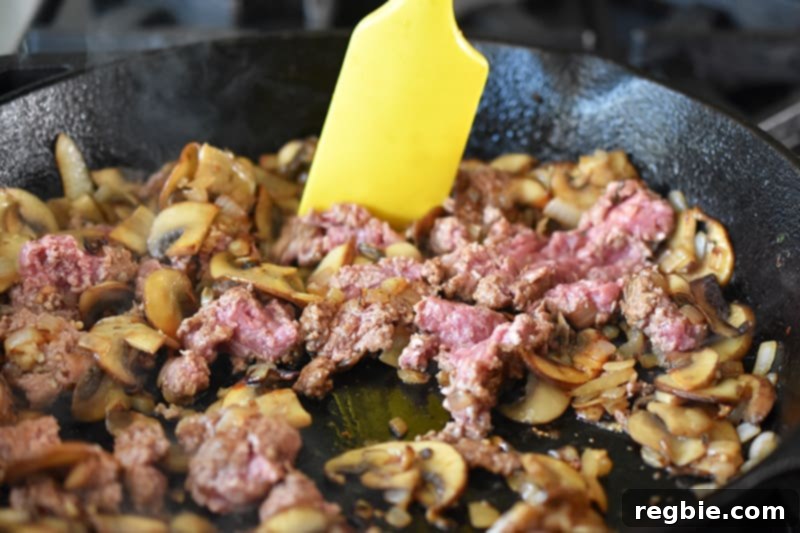 Ground beef is stirred into the pan with fragrant mushrooms, onions, and minced garlic, preparing to cook thoroughly.