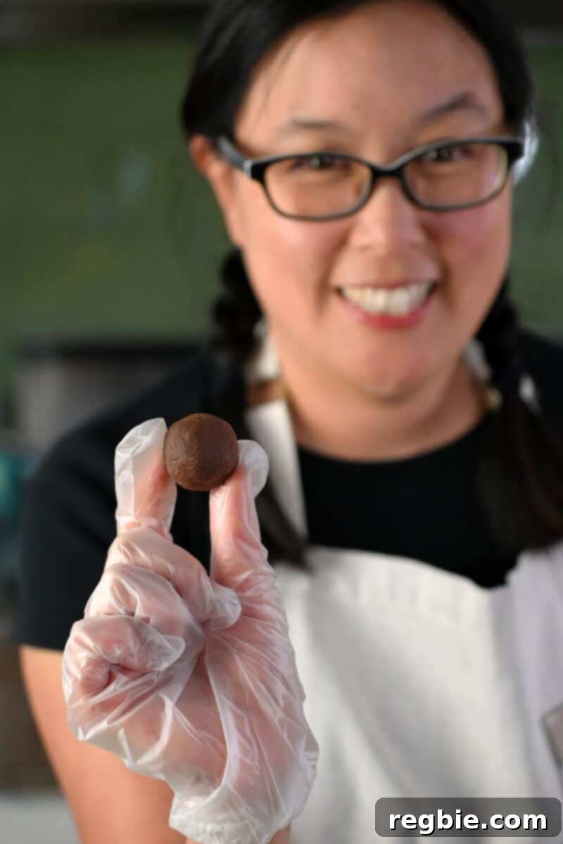 A smiling Asian woman in glasses is holding a round chocolate truffle in her gloved hand, demonstrating the final rolling step.