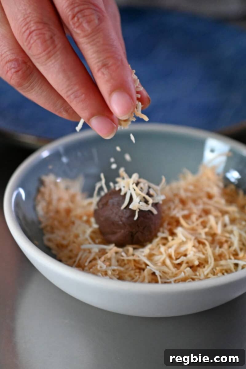 A hand is sprinkling toasted shredded coconut onto a paleo and vegan chocolate truffle in a small bowl, showing the final coating step before serving.