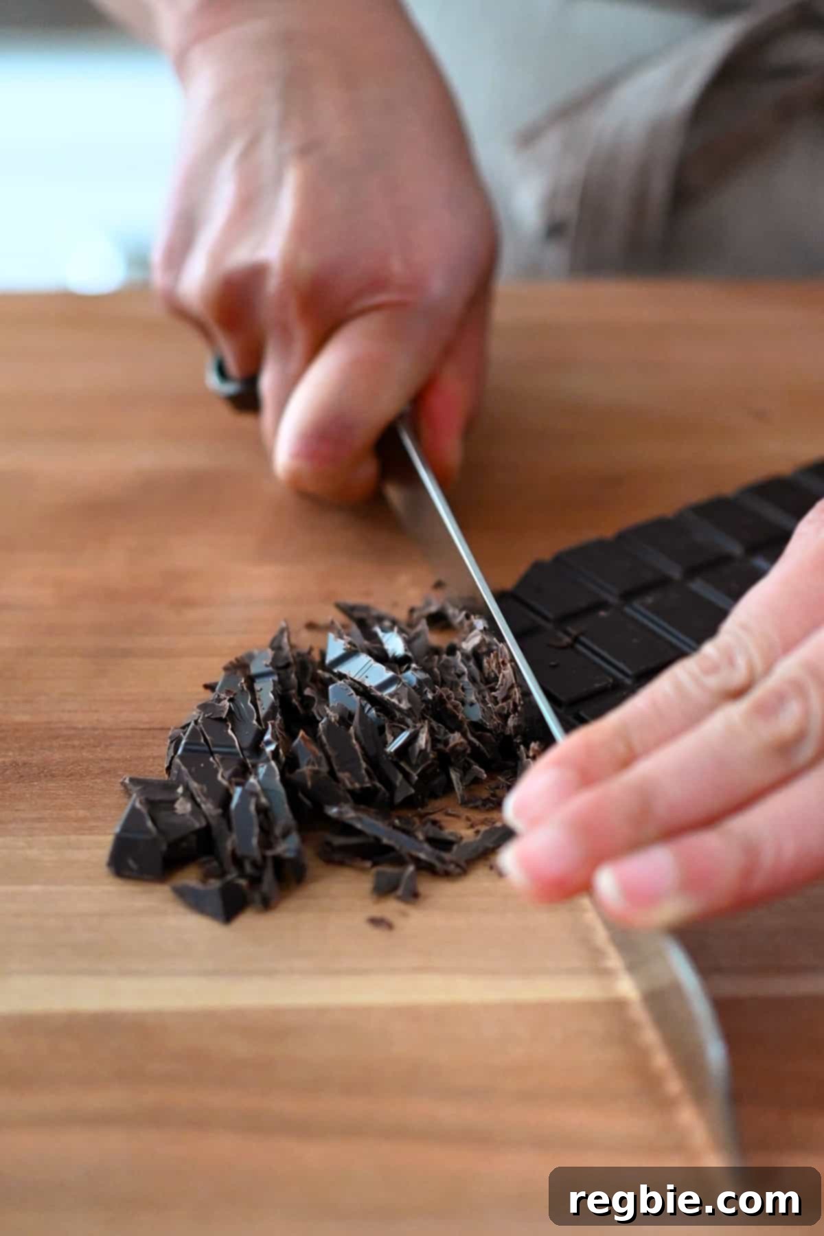 A closeup of thinly slicing a bar of dark chocolate with a chef's knife, demonstrating the proper technique for preparing chocolate for ganache.
