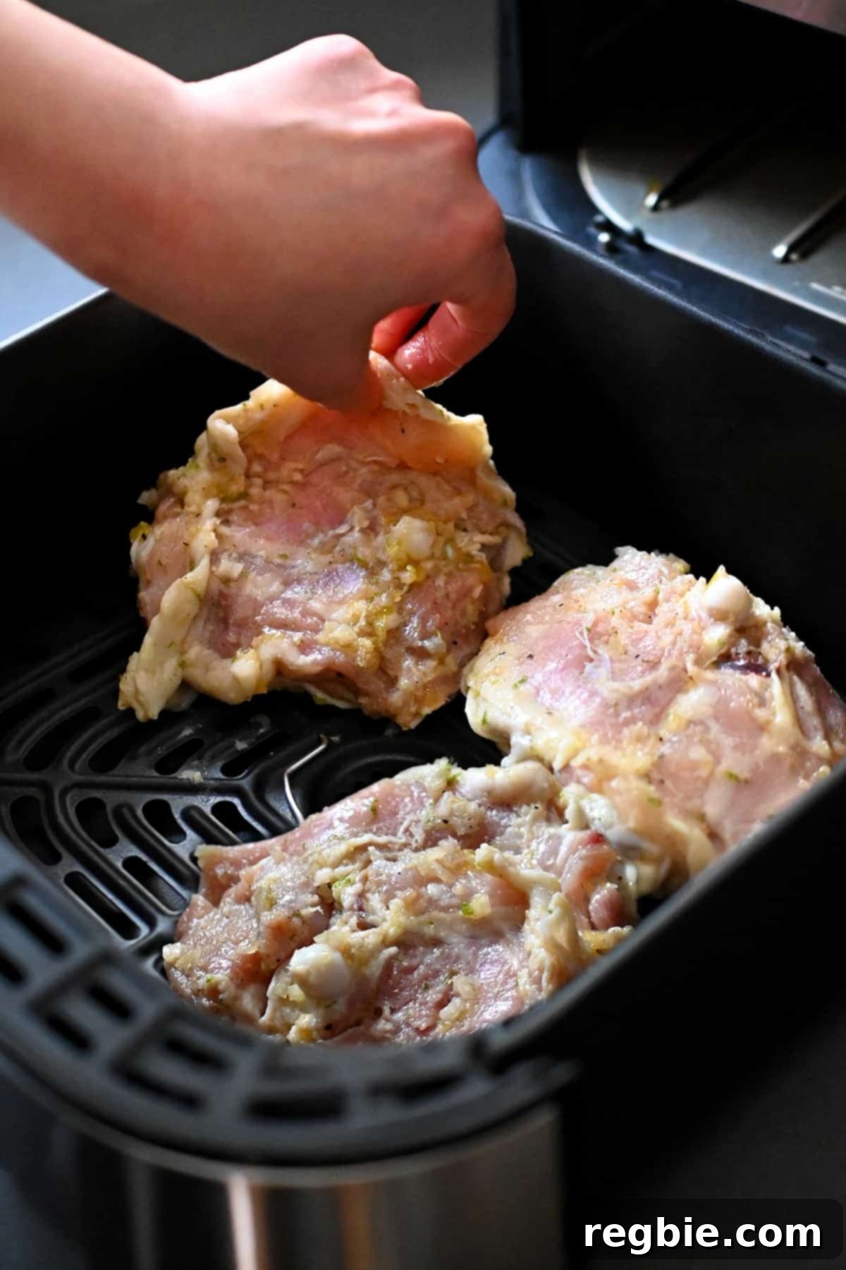 A hand is placing raw lemongrass chicken thighs, skin side down, into an open air fryer basket.