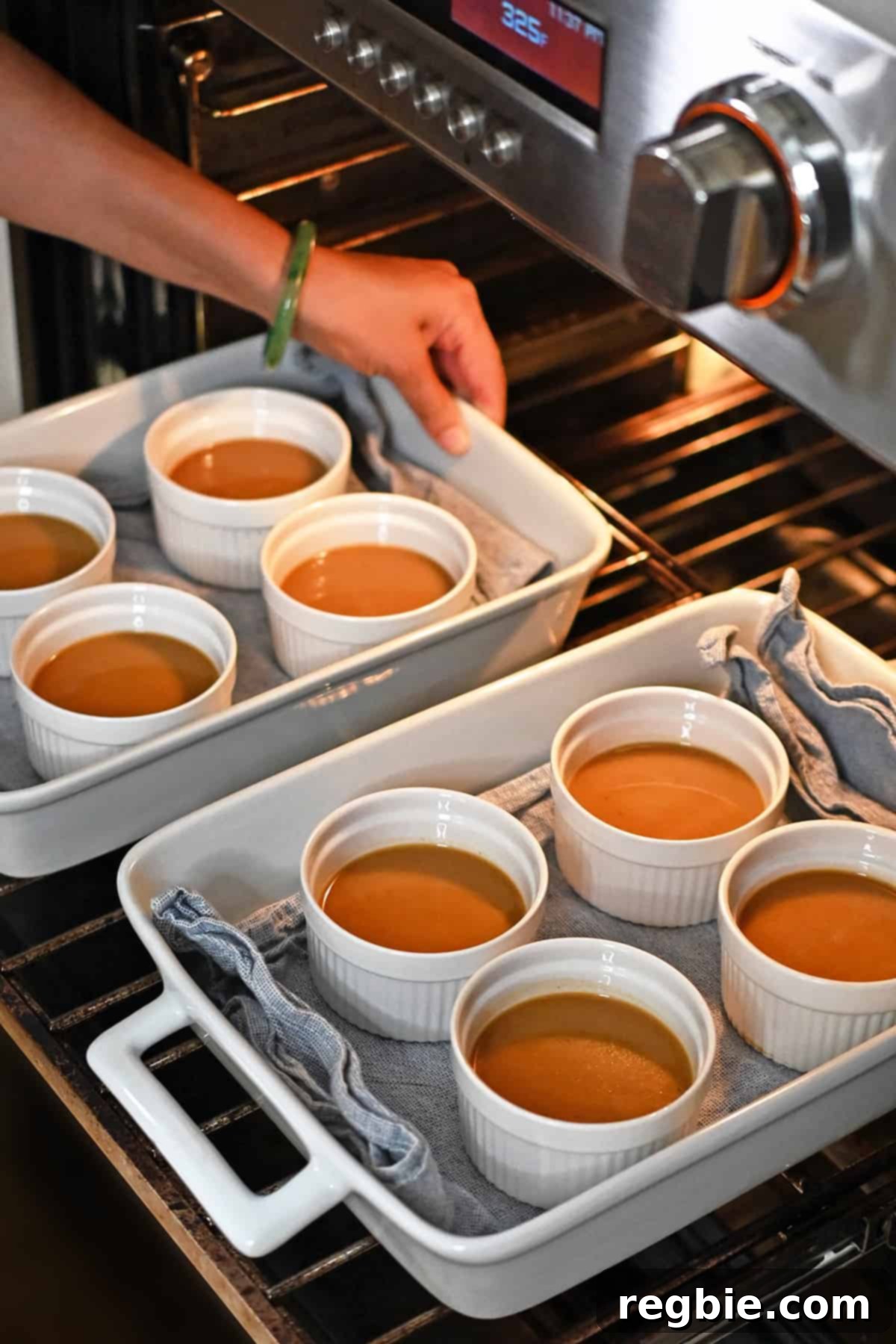 Decadent Paleo Pumpkin Custard 11 Two white casserole dishes, each holding four ramekins of pumpkin custard, are positioned inside an open oven, ready for the water bath.