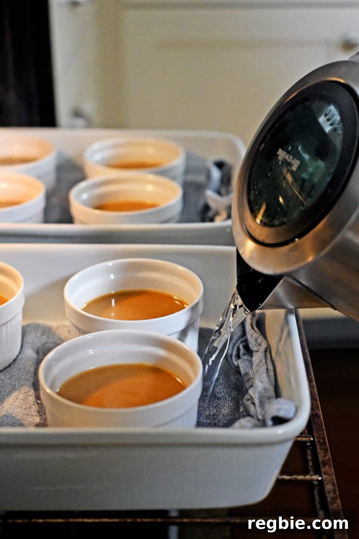 Decadent Paleo Pumpkin Custard 12 Boiling water is being poured from a kettle into a casserole dish containing ramekins of pumpkin custard, which is already in an open oven, setting up the water bath.