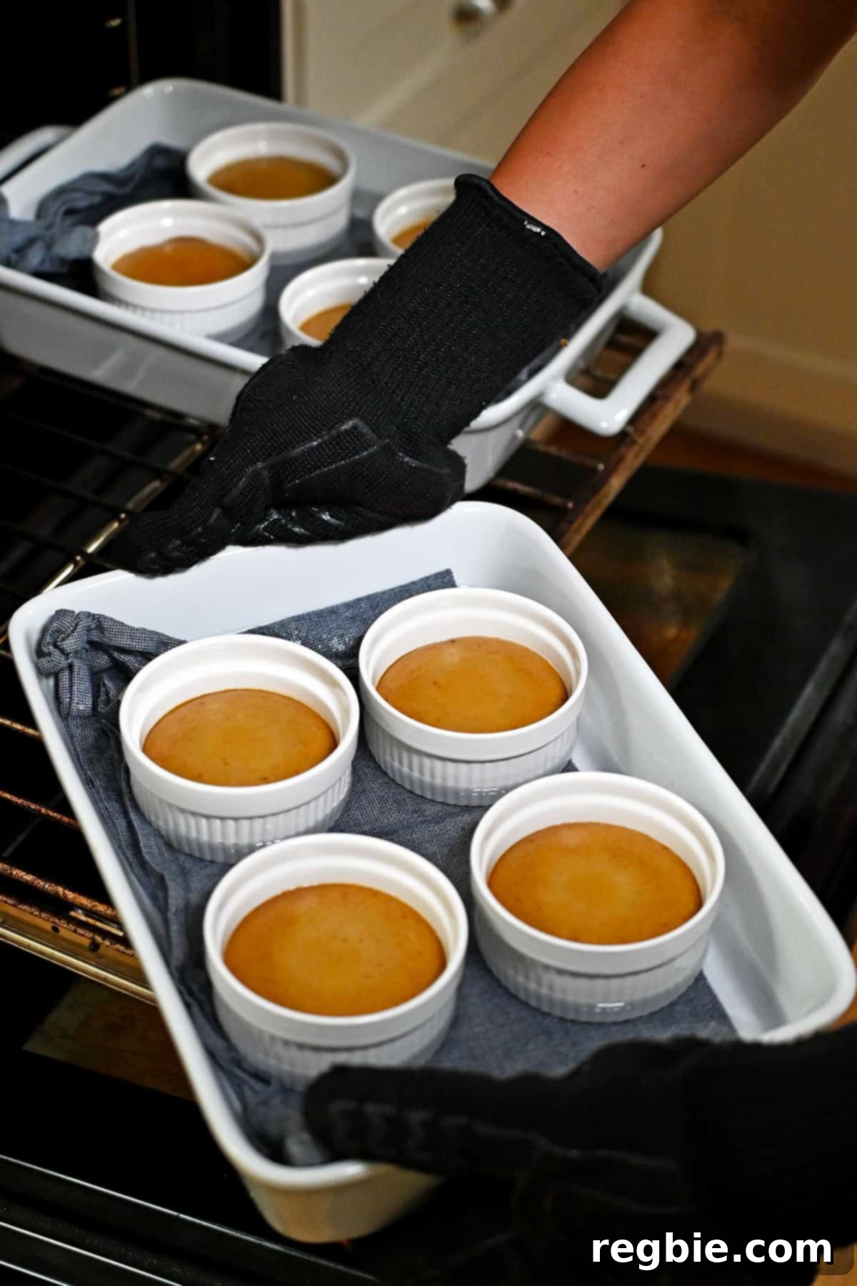 Decadent Paleo Pumpkin Custard 13 A hand is carefully taking a casserole pan filled with four perfectly cooked pumpkin custards out of the oven, steam gently rising.