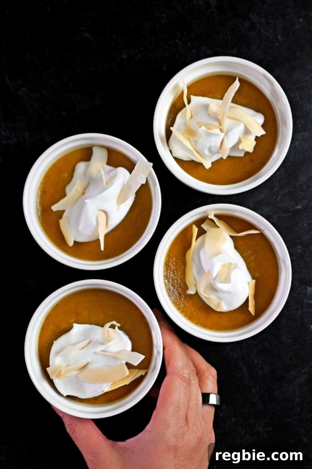 Decadent Paleo Pumpkin Custard 16 An overhead shot of four perfectly set pumpkin custards in ramekins, with a hand reaching in to grab one, highlighting their inviting presentation.