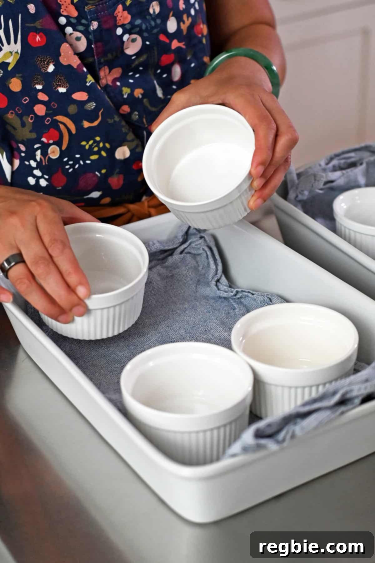 Decadent Paleo Pumpkin Custard 4 A hand is gently placing a white ramekin into a casserole dish lined with a blue and white striped cloth, preparing for the water bath.
