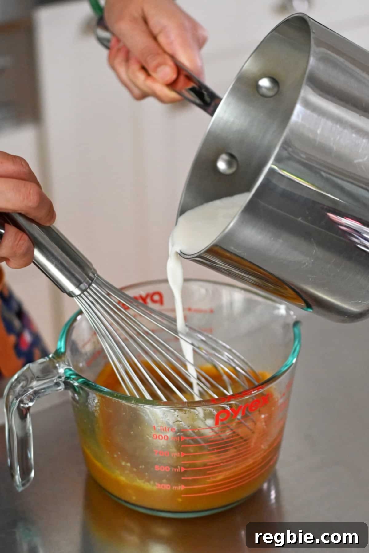 Decadent Paleo Pumpkin Custard 7 A stream of hot coconut milk is being poured into a large measuring cup containing whisked eggs and maple syrup, illustrating the tempering process.