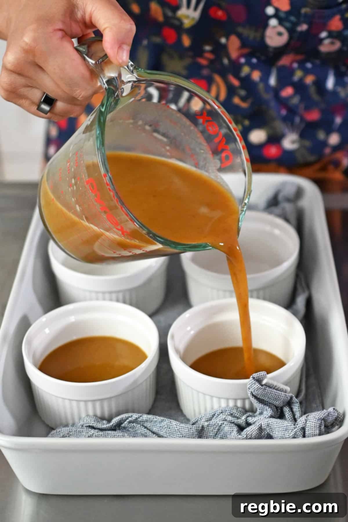 Decadent Paleo Pumpkin Custard 10 The pumpkin custard mixture is being carefully poured into white ramekins arranged in a casserole dish lined with a blue and white towel, ready for baking.
