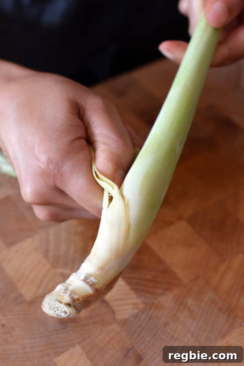 Lemongrass Cooking Guide 3 A person peeling off the tough outer leaves of a lemongrass stalk over a wooden cutting board