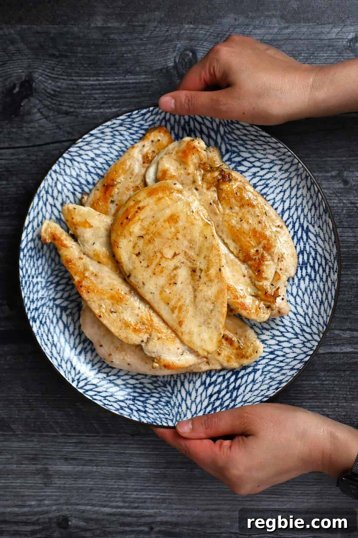 How I Cook Juicy, Tender Chicken Breasts Every Time 2 Two hands are holding a blue plate filled with golden brown, perfectly cooked boneless, skinless chicken breasts.