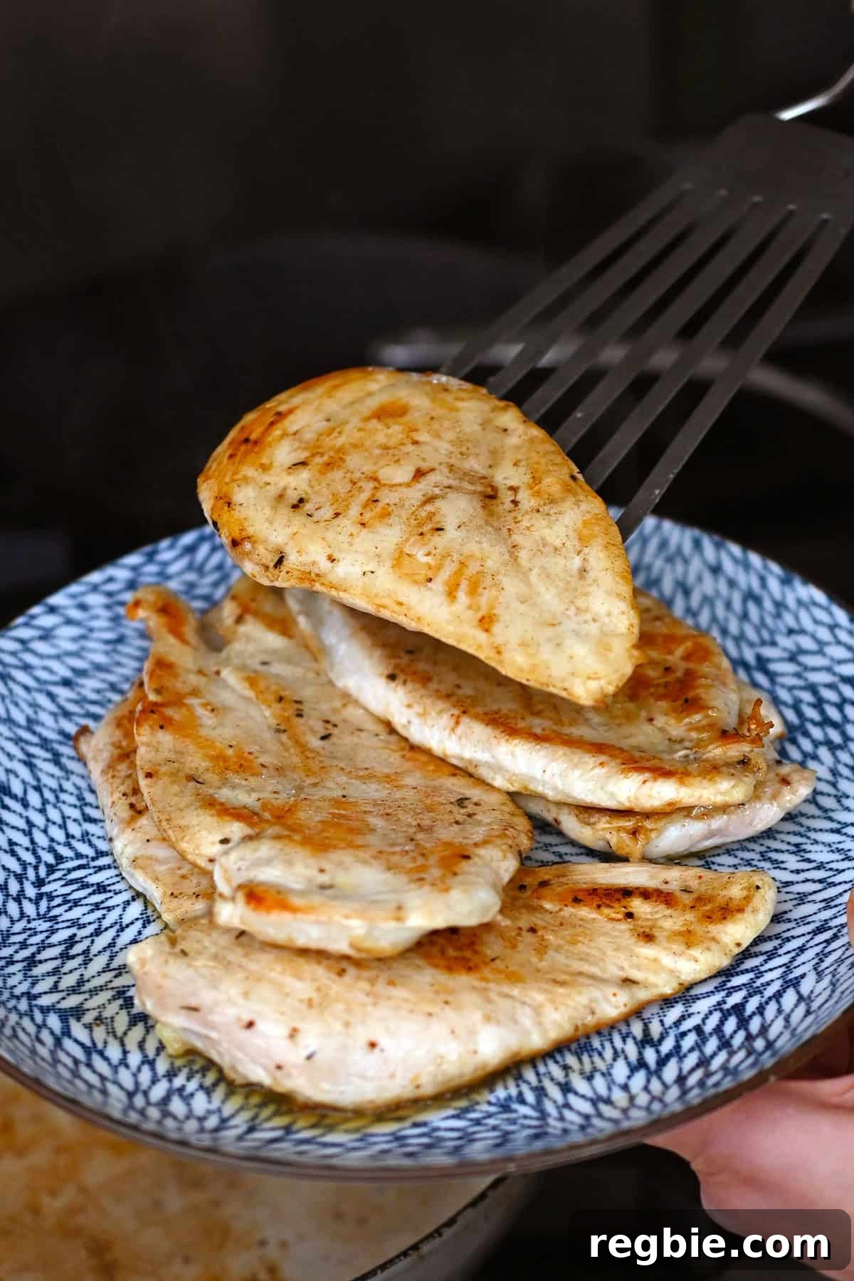 How I Cook Juicy, Tender Chicken Breasts Every Time 11 A person is using a metal fish spatula to transfer a golden brown chicken breast from a skillet onto a blue plate with other cooked chicken breasts.