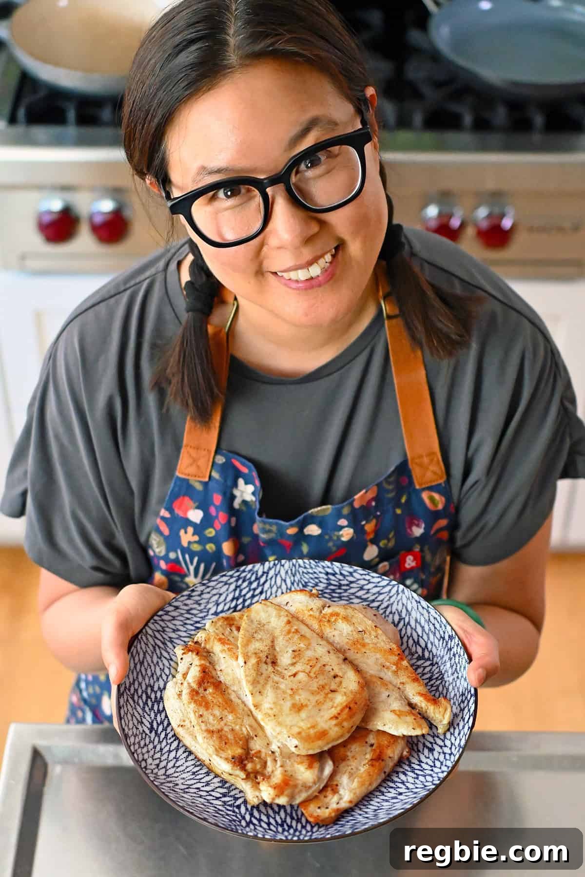 How I Cook Juicy, Tender Chicken Breasts Every Time 3 A smiling Asian woman in pigtails and black glasses is holding a blue plate filled with perfectly cooked boneless skinless chicken breasts.