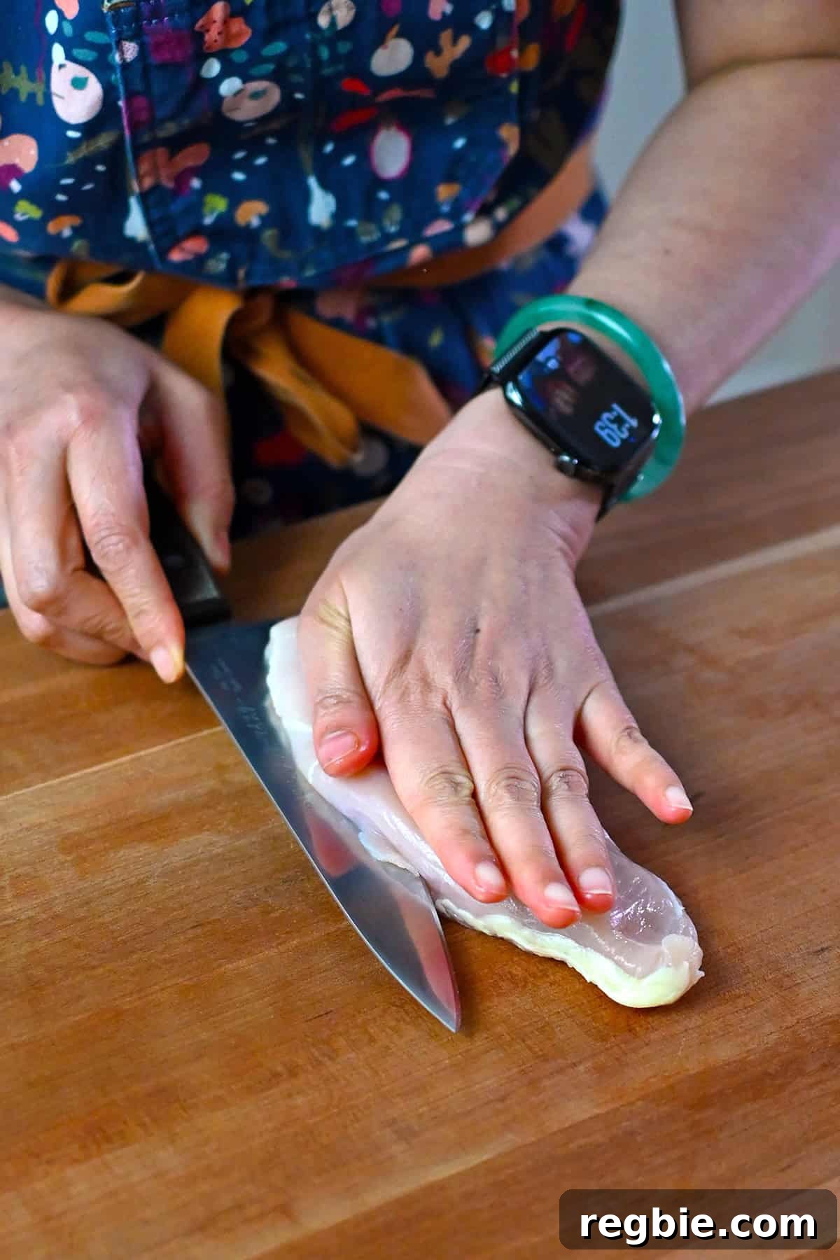 How I Cook Juicy, Tender Chicken Breasts Every Time 5 A person's hand is resting flat on top of a raw boneless skinless chicken breast as they use their other hand to hold a chef's knife to slice the breast in half horizontally.